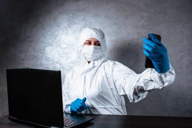 Beyaz maskeli ve tıbbi eldivenli viral koruyucu giysili genç bir kadın görüntülü bir aramada arkadaşlarıyla konuşuyor çünkü doktor koronavirüs salgınının yayılması nedeniyle karantinada..