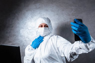 Beyaz maskeli ve tıbbi eldivenli viral koruyucu giysili genç bir kadın görüntülü bir aramada arkadaşlarıyla konuşuyor çünkü doktor koronavirüs salgınının yayılması nedeniyle karantinada..