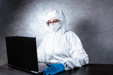 Beyaz maske takan ve beyaz koruyucu giysili tıbbi eldiven giyen bir virüs uzmanı dizüstü bilgisayarla çalışıyor ve koronavirüs hakkında bilgi topluyor. İzole edilmiş ve salgın yüzünden panik içinde..