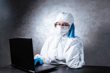 Beyaz maske takan ve beyaz koruyucu giysili tıbbi eldiven giyen bir virüs uzmanı dizüstü bilgisayarla çalışıyor ve koronavirüs hakkında bilgi topluyor. İzole edilmiş ve salgın yüzünden panik içinde..