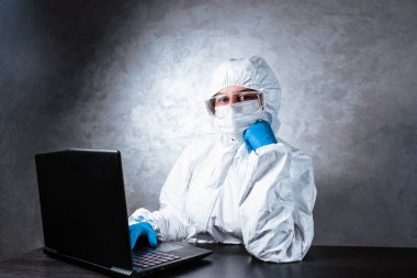 Beyaz maske takan ve beyaz koruyucu giysili tıbbi eldiven giyen bir virüs uzmanı dizüstü bilgisayarla çalışıyor ve koronavirüs hakkında bilgi topluyor. İzole edilmiş ve salgın yüzünden panik içinde..