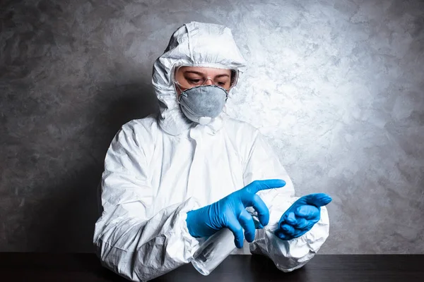 A nurse in a medical face mask and viral protective suit. The doctor is sitting at the table in a medical face mask and a viral protective suit. The doctor disinfects the hands.