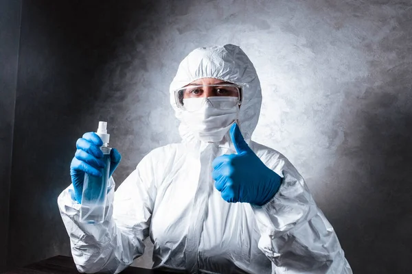 Nurse in a medical face mask and a viral protective suit. The doctor is sitting at the table in a medical face mask and a viral protective suit. The doctor disinfects the hands and shows the class.