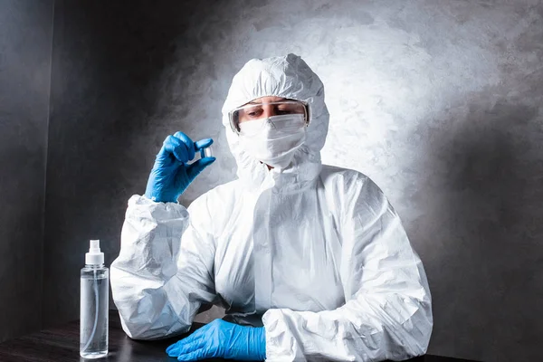 Nurse in a medical face mask and a viral protective suit. The doctor is sitting at the table in a medical face mask and a viral protective suit. The doctor holds a pill in his hands.