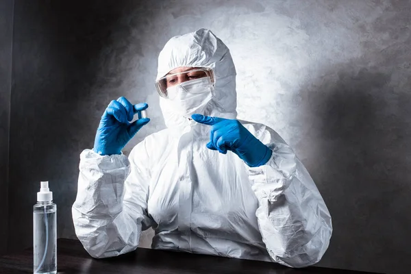Nurse in a medical face mask and a viral protective suit. The doctor is sitting at the table in a medical face mask and a viral protective suit. The doctor holds a pill in his hands.