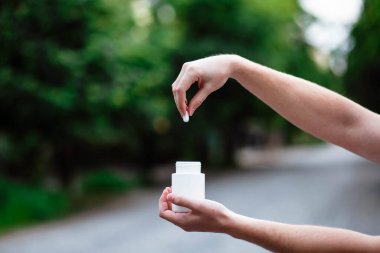 Adamın elinde ağaçların arkasına izole edilmiş haplarla dolu bir ilaç şişesi var. Yaklaştın. Yüksek çözünürlüklü. Elleri beyaz plastik bir şişe tutuyor..