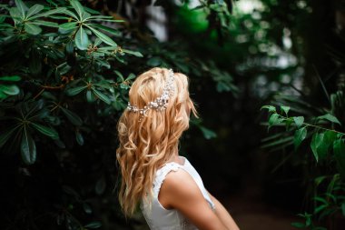 Yeşilliğin arka planında gelinlik içinde duran çekici sarışın gelin portresi. Botanik yeşili bahçesinde bir kadın var. Kıvırcık sarı saçlı güzel genç kadınlar.