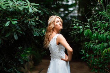 Yeşillik arka planında gelinlik giymiş çekici sarışın bir gelinin portresi. Botanik yeşili bahçesindeki gelin. Kıvırcık sarı saçlı güzel bir genç kadın. Evlilik..