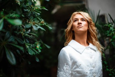 Yeşillik arka planında gelinlik giymiş çekici sarışın bir gelinin portresi. Botanik yeşili bahçesindeki gelin. Kıvırcık sarı saçlı güzel bir genç kadın. Evlilik..