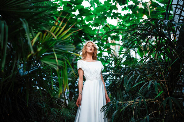Portrait of an attractive blonde bride standing in a wedding dress on a background of greenery. The bride in the botanical green garden. Beautiful young woman with curly blond hair. Marriage.