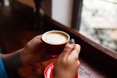 Genç bir kızın ellerinde bir bardak kapuçino. Ahşap zemin üzerinde kahve, el planı ve kahve olan kırmızı bardağı tutan bir kız. Üst Manzara. Mesaj için yer. Sütlü kapuçino..