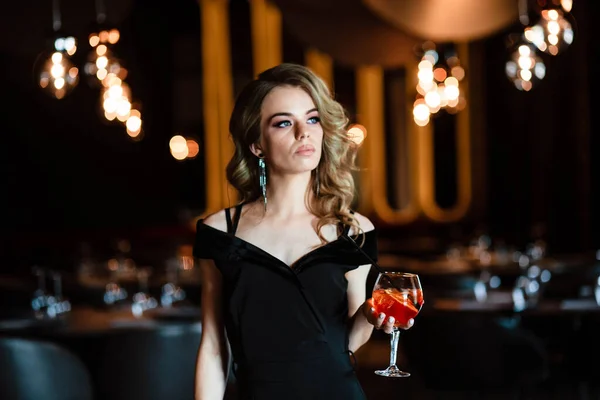 Güzel sarışın, genç bir kız aperol spreyi tutuyor. Bardakta kokteyl aperol spreyi. Restorandaki genç bir kızın portresi. Bir kızın portresi. Bardakla restoranda genç bir kız..