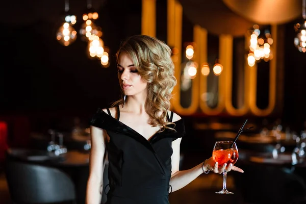 Güzel sarışın, genç bir kız aperol spreyi tutuyor. Bardakta kokteyl aperol spreyi. Restorandaki genç bir kızın portresi. Bir kızın portresi. Bardakla restoranda genç bir kız..