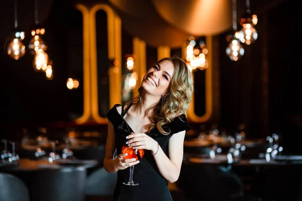 Güzel sarışın, genç bir kız aperol spreyi tutuyor. Bardakta kokteyl aperol spreyi. Restorandaki genç bir kızın portresi. Bir kızın portresi. Bardakla restoranda genç bir kız..