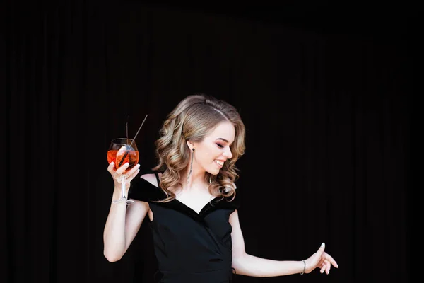 Güzel sarışın, genç bir kız aperol spreyi tutuyor. Bardakta Aperol kokteyli şırıngası. Restorandaki genç bir kızın portresi. Siyah arka planı olan bir restoranda genç bir kız..