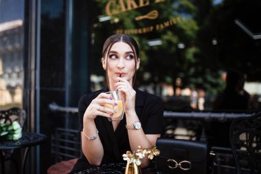 Şehirdeki bir kızın portresi. Siyah elbiseli kız. Genç ve güzel bir kadın. Makyaj. Kız elinde bir limonata tutuyor ve onu içiyor. Bir kadının ellerinde limonata.