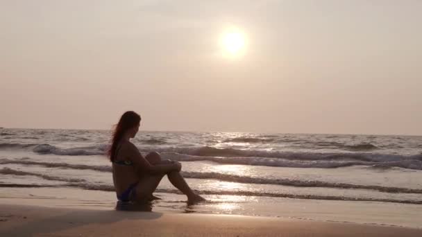 Νεαρή λεπτή κοπέλα σε ένα κοστούμι λουσίματος κάθεται στην παραλία το ηλιοβασίλεμα. 4k — Αρχείο Βίντεο