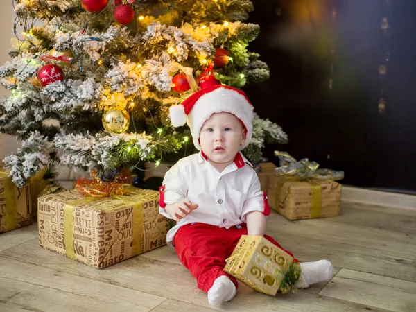 Noel ağacı, hediyeler ile Santas kırmızı şapkalı küçük güzel bebek oturur — Stok fotoğraf