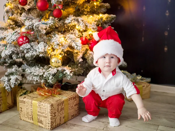 Işıklar ve evde topları ile zarif bir Noel ağacı altında kırmızı Santa şapka içinde küçük bir çocuk — Stok fotoğraf