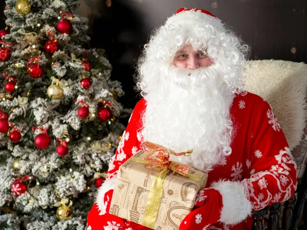 Santa con una barba blanca y con un regalo en sus manos sobre el fondo del árbol de Navidad en un interior festivo — Foto de Stock