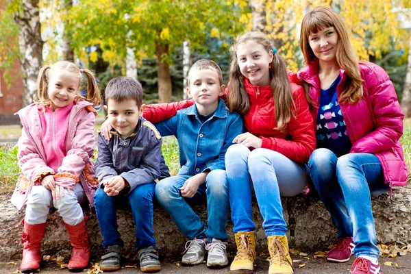 Los niños alegres y la mujer joven en las chaquetas brillantes a la moda en el parque otoñal por la tarde — Foto de Stock