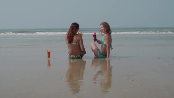 Két vidám szép fiatal lány ül a hátukon a strand, a tenger mellett. 4k