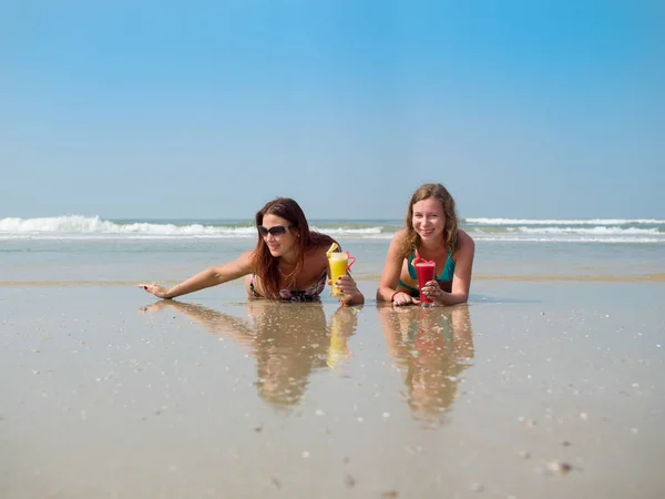 Deux filles blanches avec un verre de jus de fruits dans leurs mains sont couchées dans le sable sur la plage — Photo