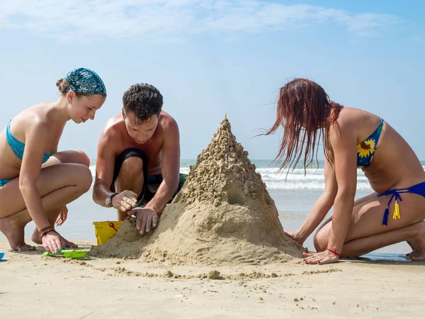 Ett företag av unga människor bygger ett sandslott på stranden på en hård dag. — Stockfoto