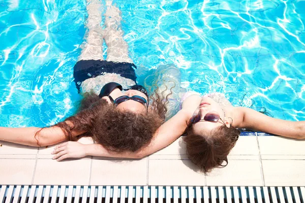 Dos hermosas chicas jóvenes en gafas de sol están tumbados en el borde de la piscina relajante — Foto de Stock