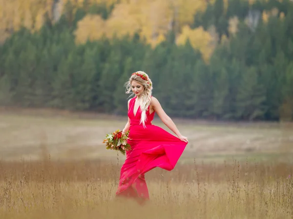 Mladá dívka v červených dlouhých šatech s účesu a s podzimním buketkou rozvine lem šatů v přírodě — Stock fotografie