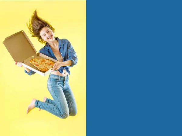 Smiling young girl or girl in jeans and a blue shirt is having fun jumping high while holding a pizza box in hands on a yellow background — Stock Photo, Image
