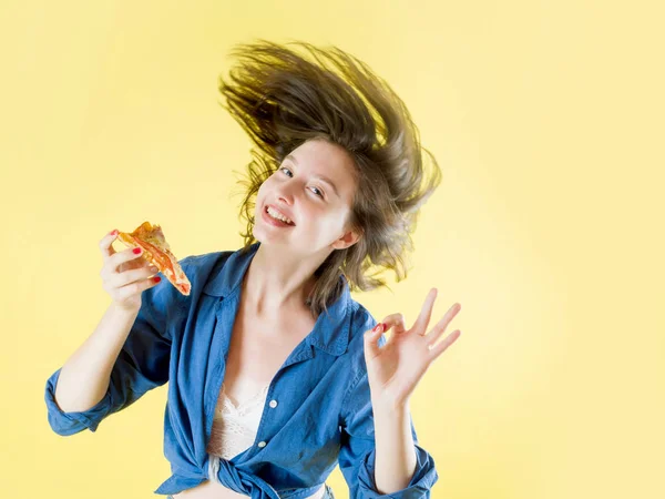 Chica con un pedazo de pizza en las manos con el pelo volando sobre un fondo amarillo —  Fotos de Stock