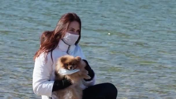 Spitz meisje en hond in beschermende chirurgische masker op de natuur op een lentedag. Coronavirus COVID-19 is gevaarlijk voor huisdieren. Red Spitz met een medisch masker. Coronavirus Pandemische voorzorgsmaatregelen — Stockvideo