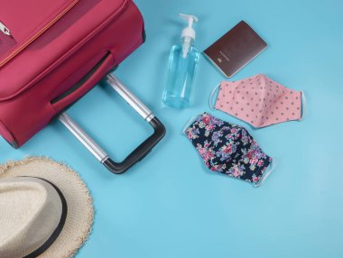 Yolculuk sırasında COVID-19 önleme ve yeni normal yaşam tarzı konsepti. İki renkli kumaş yüz maskesi, alkol el dezenfektanı, şapka, bavul ve mavi arka planda pasaport..