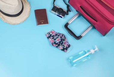Yolculuk sırasında COVID-19 önleme ve yeni normal yaşam tarzı konsepti. Çiçek desenli kumaş maskeler, alkol dezenfektanı jeli ve seyahat aksesuarları mavi arka planda fotokopi alanı ile.