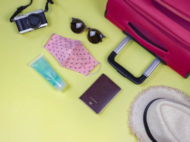 Yolculuk sırasında COVID-19 önleme ve yeni normal yaşam tarzı konsepti. Pembe benekli kumaş maskeler, alkol dezenfektanı jeli ve sarı arka plandaki seyahat aksesuarları..