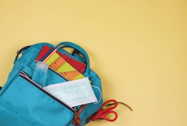 Okula geri dönerken COVID-19 önleme ve yeni normal yaşam tarzı kavramı. Okul malzemeleriyle dolu sırt çantası, cerrahi maske ve alkol dezenfektanı sarı arka planda fotokopi alanı olan el jeli..