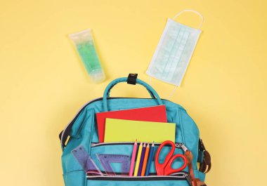 Okula geri dönerken COVID-19 önleme ve yeni normal kavram. Okul malzemeleriyle dolu sırt çantasının üst görüntüsü, ameliyat maskesi ve sarı arka planda fotokopi alanı olan alkol temizleyici jel..