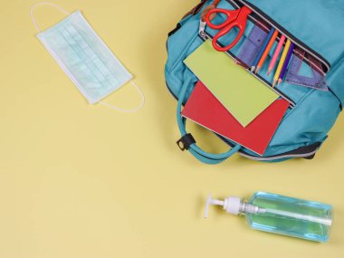 Okula geri dönerken COVID-19 önleme ve yeni normal kavram. Okul malzemeleriyle dolu sırt çantasının üst görüntüsü, ameliyat maskesi ve sarı arka planda fotokopi alanı olan alkol temizleyici jel..