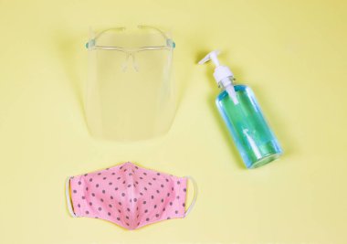 Coronavirus veya COVID-19 önleme ve yeni normal yaşam tarzı kavramı. Yüzün üst görüntüsü titrek, alkol el dezenfektanı jeli ve sarı arka planda pembe puantiyeli kumaş maskesi..