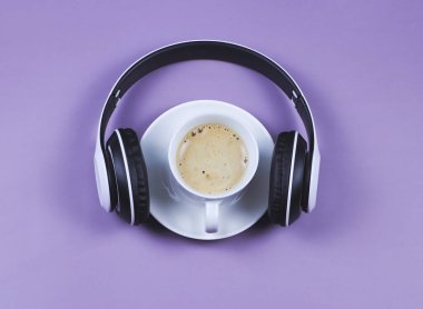 Üst ya da düz kulaklıklar ve mor zemin üzerinde kahve fincanı. Müzik ve kahve rahatlatıcı zaman kavramı.