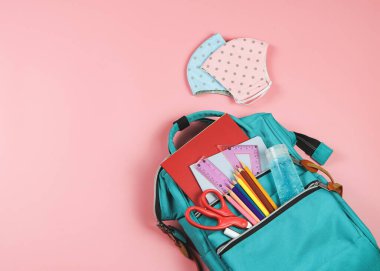 Okula geri dönerken COVID-19 önleme ve yeni normal kavram. Okul malzemeleriyle dolu sırt çantası, pembe ve mavi benekli kumaş maskeler ve pembe arka planda dezenfektan jeli üst görünüm.