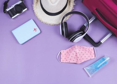 Yolculuk sırasında COVID-19 önleme ve yeni normal yaşam tarzı konsepti. Pembe benekli kumaş maskeler, alkol dezenfektanı jeli ve mor arkaplanlı seyahat aksesuarları..