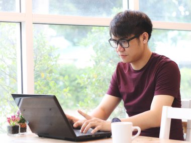 Kahve dükkanında oturan gözlüklü Asyalı genç adam masadaki bilgisayar ekranına dikkatlice bakıyor. , 