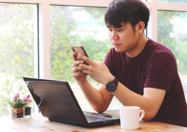 Kahve dükkanının penceresinde oturan Asyalı genç adam masada bilgisayar laptopuyla cep telefonuyla yazı yazıyor..