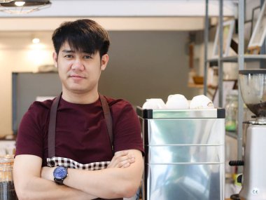 Asyalı genç adam kahve makinesinin önünde duruyor, kendi kahve dükkanında, kollarını kavuşturup kameraya bakıyor..