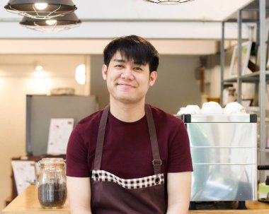 Asyalı genç adam kahve makinesinin önünde oturmuş kameraya gülümsüyor..