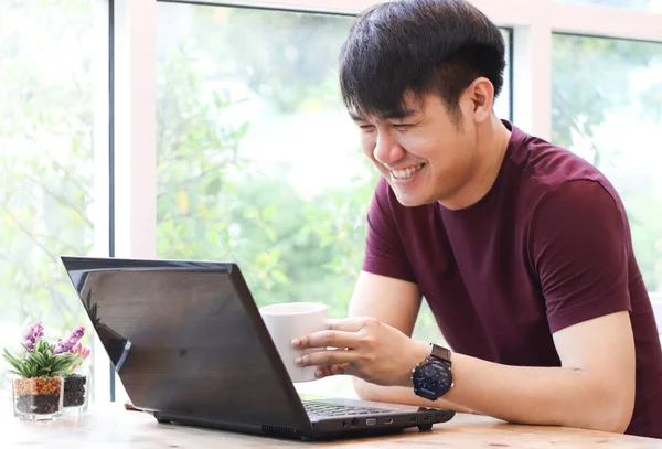 Asian young man sitting by the window in coffee shop with computer laptop on the table , looking at computer screen and laughing.