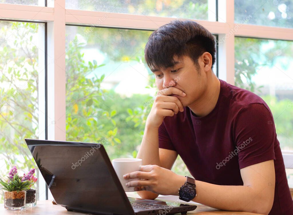Asian young man working in coffee shop drinking coffee , looking at his computer laptop on the table and touching his mouth thoughtfully.