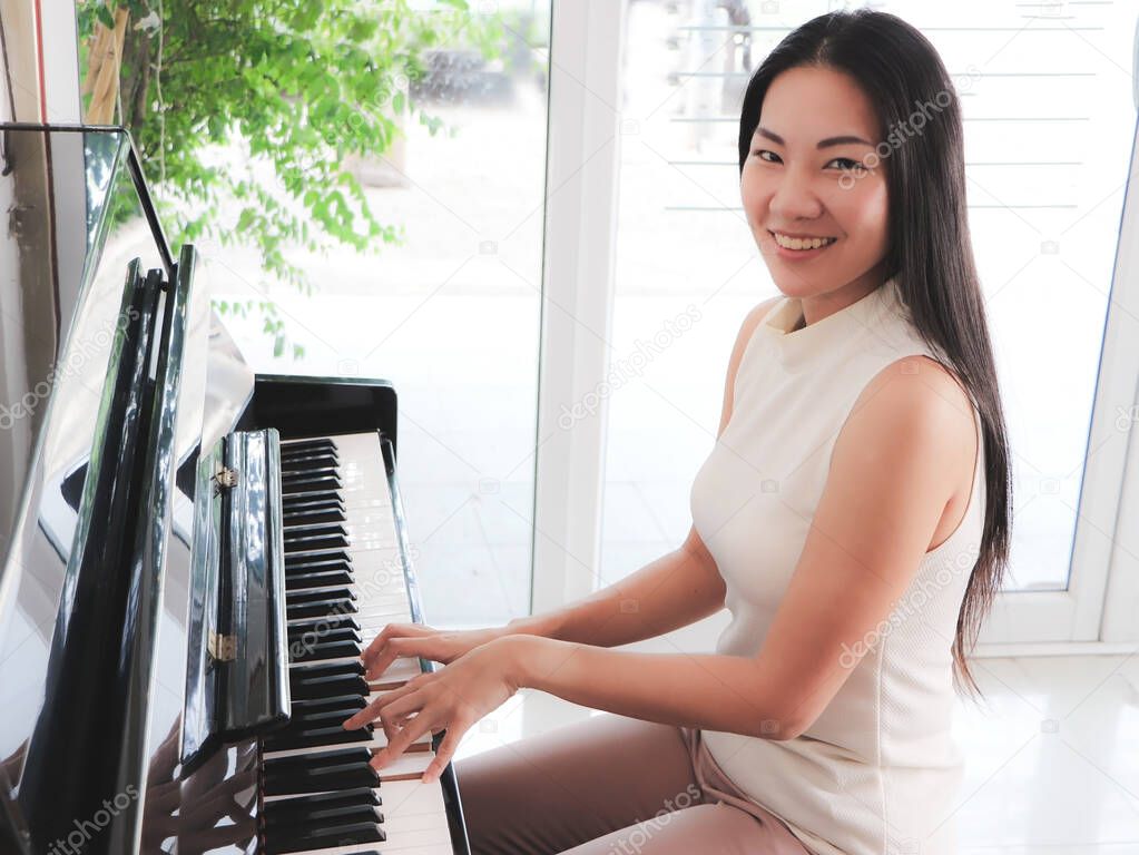 Asian young woman playing pinao upright in the white room and smiling at the camera.